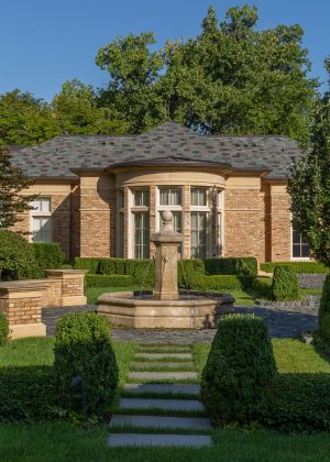 Light brick home with large fountain and brick driveway, bordered by boxwood hedges and white hydrangeas.