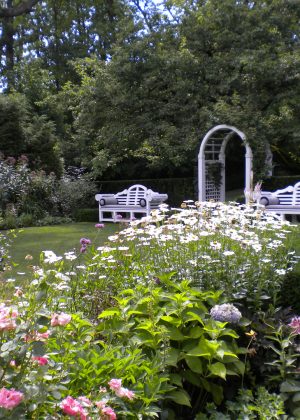 Serene sitting area with white benches and arbor nestled in perennial garden with pink and white flowers