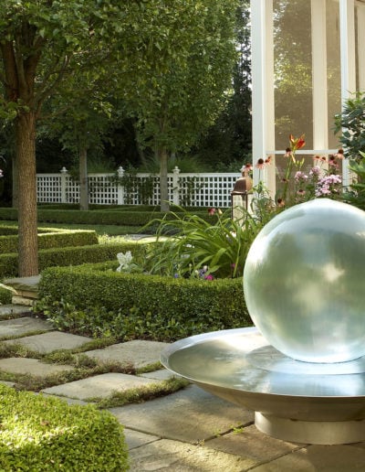 «Un jardín tranquilo en las afueras de Chicago, con un porche acristalado rodeado de setos bajos, flores y una fuente con una esfera transparente sobre una superficie pavimentada».