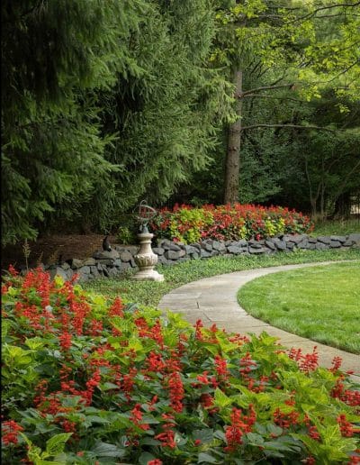 El sendero del jardín serpentea a través de un parterre de flores rojas con un borde de piedra y árboles de hoja perenne al fondo.